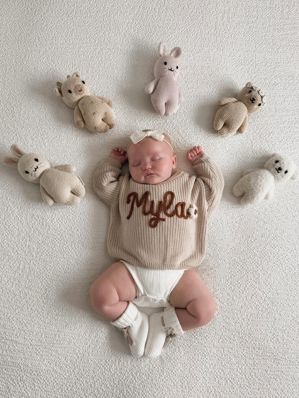 Newborn in an embroidered name sweater surrounded by soft toys