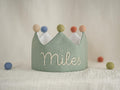 May include: A sage green fabric crown featuring the embroidered name Miles in white thread. The crown is topped with five small felt pom-poms in muted shades of beige, blue, olive green, terracotta, and peach, with a white interior lining.