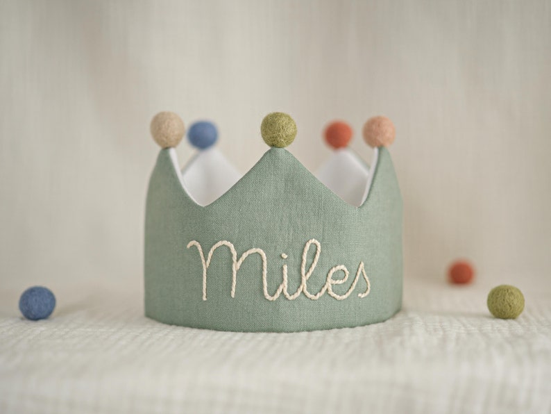 May include: A sage green fabric crown featuring the embroidered name Miles in white thread. The crown is topped with five small felt pom-poms in muted shades of beige, blue, olive green, terracotta, and peach, with a white interior lining.