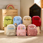 May include: A collection of seven corduroy backpacks in various colours including lime green, light blue, beige, red, cream, light pink, and dusty rose. Each backpack features a personalised name embroidered on the front and a small plush animal.
