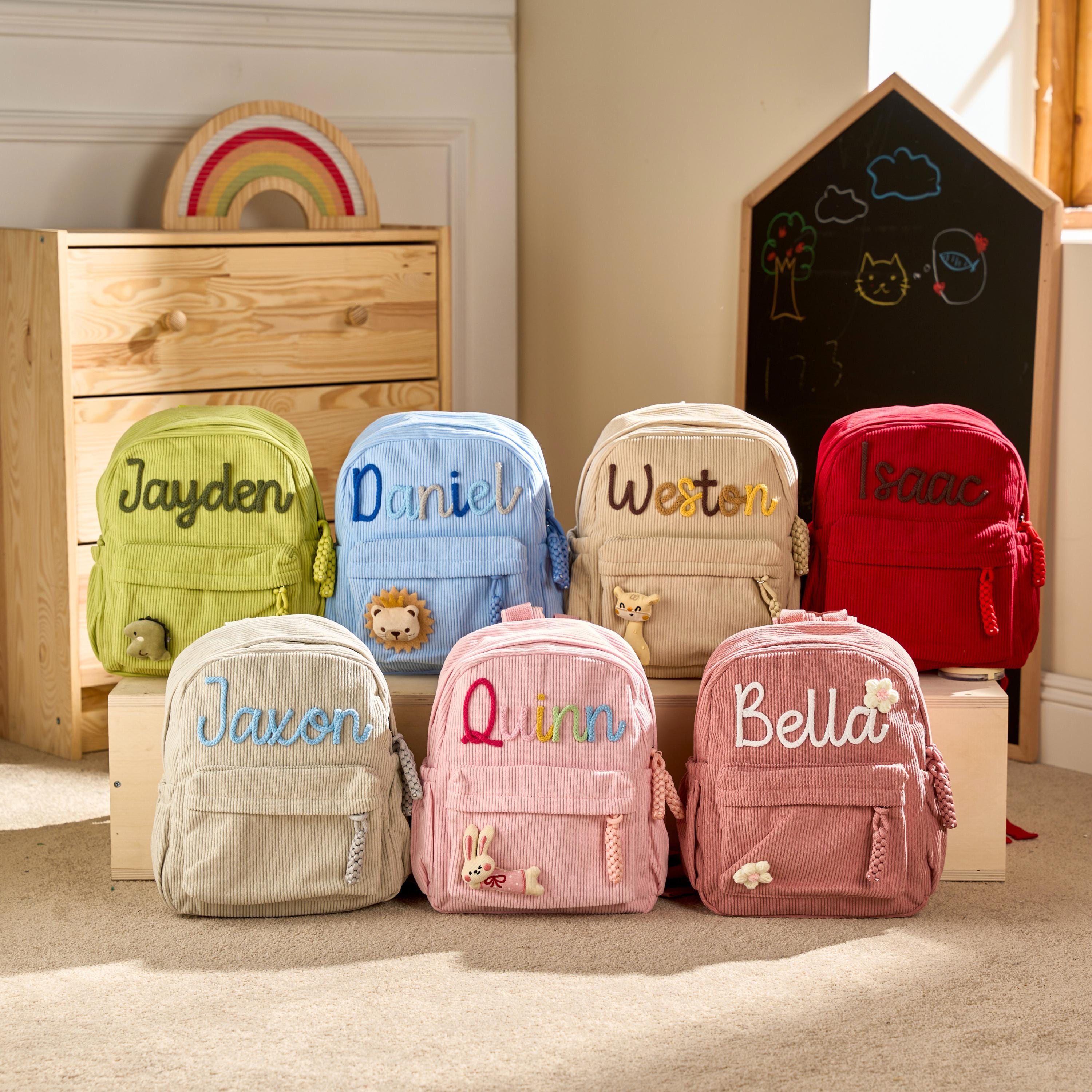 May include: A collection of seven corduroy backpacks in various colours including lime green, light blue, beige, red, cream, light pink, and dusty rose. Each backpack features a personalised name embroidered on the front and a small plush animal.