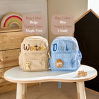May include: Two corduroy backpacks, one beige and one blue, each featuring embroidered names and small plush animal appliques. The beige bag displays the name Weston with a giraffe, while the blue bag shows the name Daniel with a lion. Text labels indicate Bag Color: Beige, Name Color: Maple Tree and Bag Color: Blue, Name Color: Cloudy.