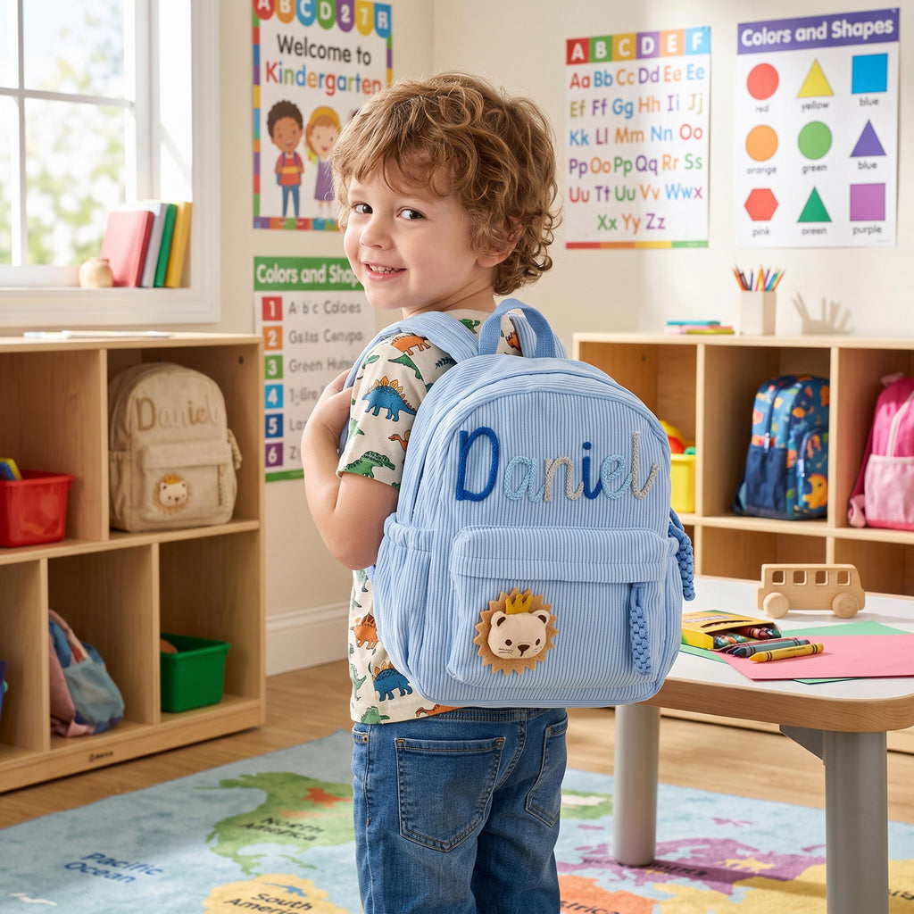 May include: A light blue corduroy backpack featuring the name Daniel embroidered in dark blue script and a small lion patch with a yellow mane on the front pocket. The backpack has adjustable shoulder straps and a top handle for easy carrying.
