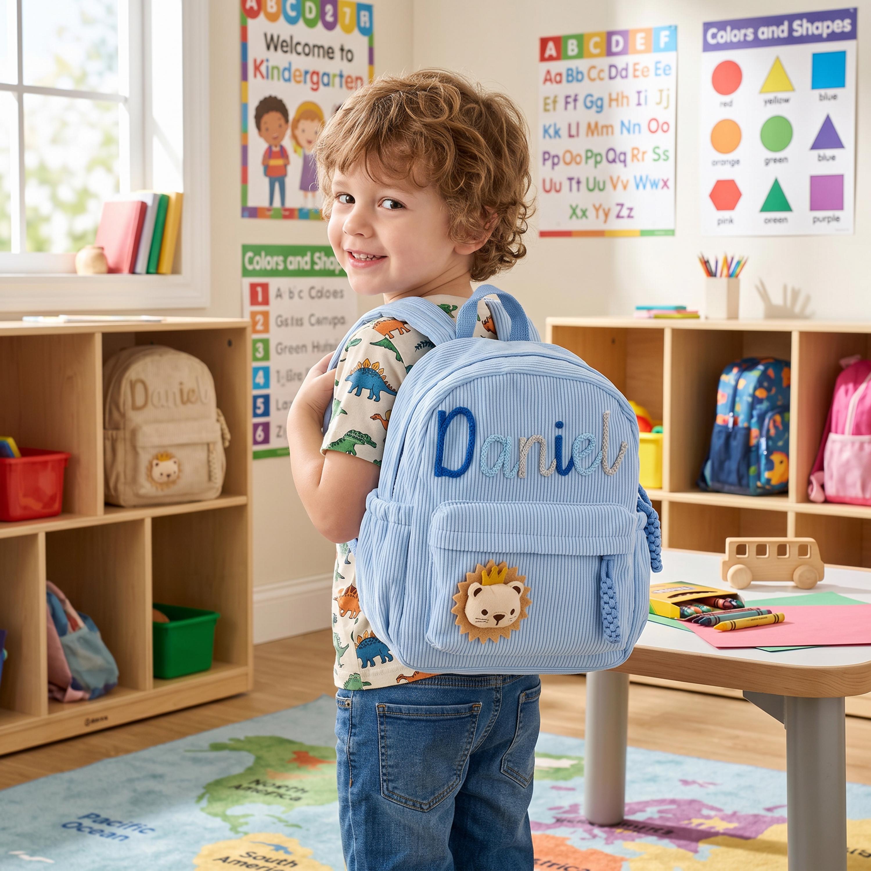 May include: A light blue corduroy backpack featuring the name Daniel embroidered in dark blue script and a small lion patch with a yellow mane on the front pocket. The backpack has adjustable shoulder straps and a top handle for easy carrying.
